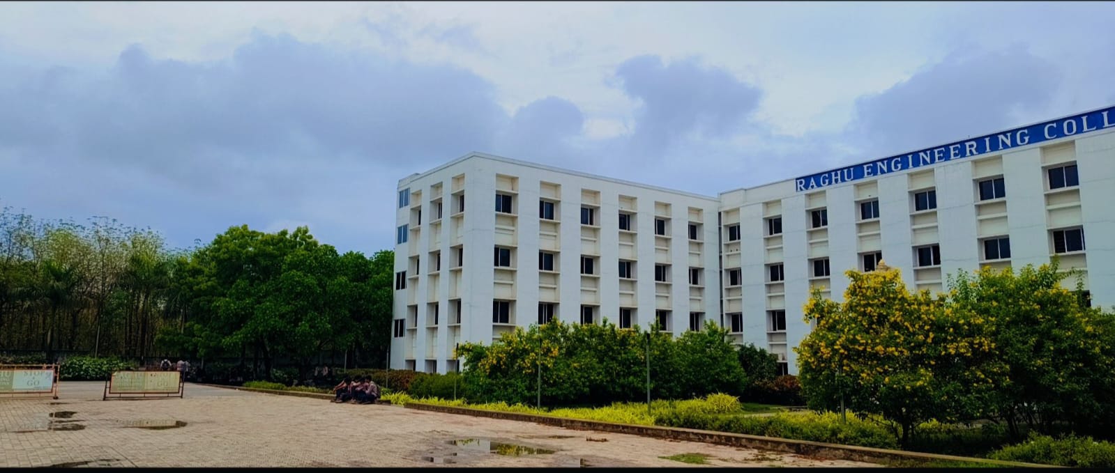 A young man stands on a sunlit street holding a camera, smiling warmly as he looks toward the lens. Lush green trees line the road, and soft afternoon light creates a peaceful, inviting atmosphere. The scene conveys a sense of joy and calm, with no visible text in the image.